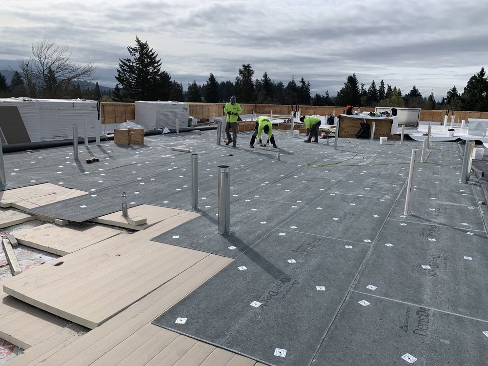 Construction workers installing roofing insulation on a flat rooftop with scattered materials and tools under overcast skies.