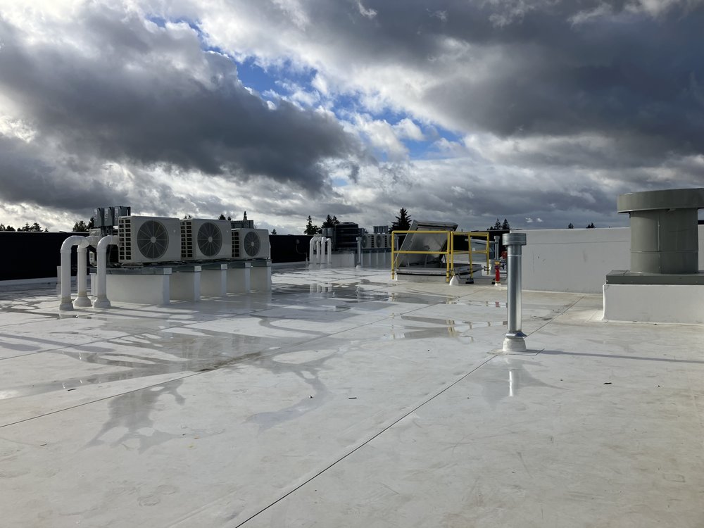 Rooftop with HVAC units and a cloudy sky in the background. The surface is wet, reflecting the equipment and clouds.