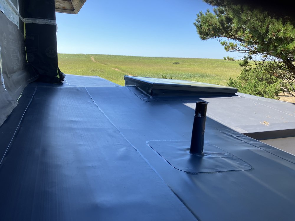 Flat rooftop with black waterproof membrane, a vent pipe, and a skylight under a clear blue sky with grassy landscape visible in the background.