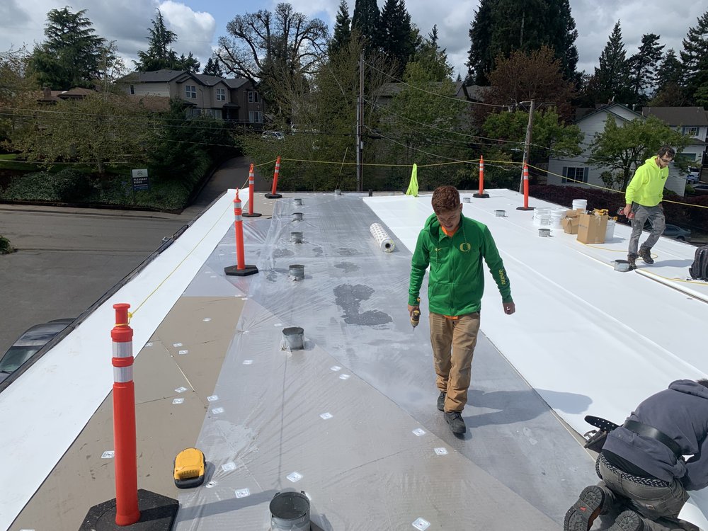 Workers installing a roof membrane on a building, with trees and houses in the background. Safety cones line the edge of the roof.