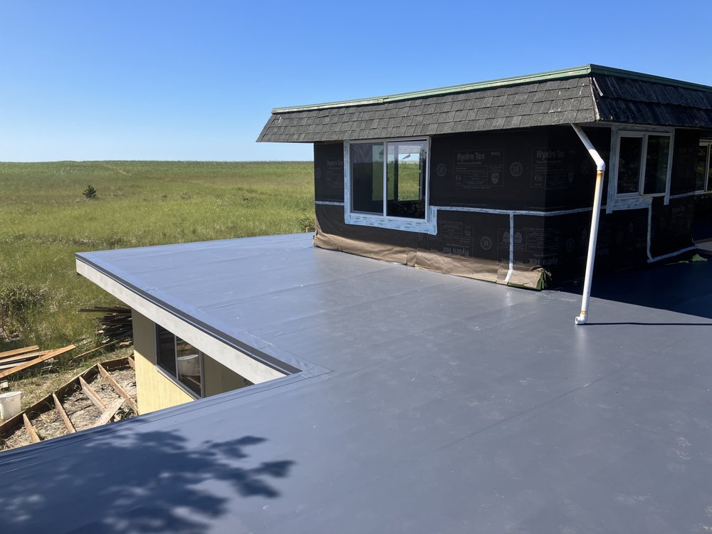 Flat rooftop with a newly installed, smooth, dark coating. A small structure with windows is partially covered in house wrap.