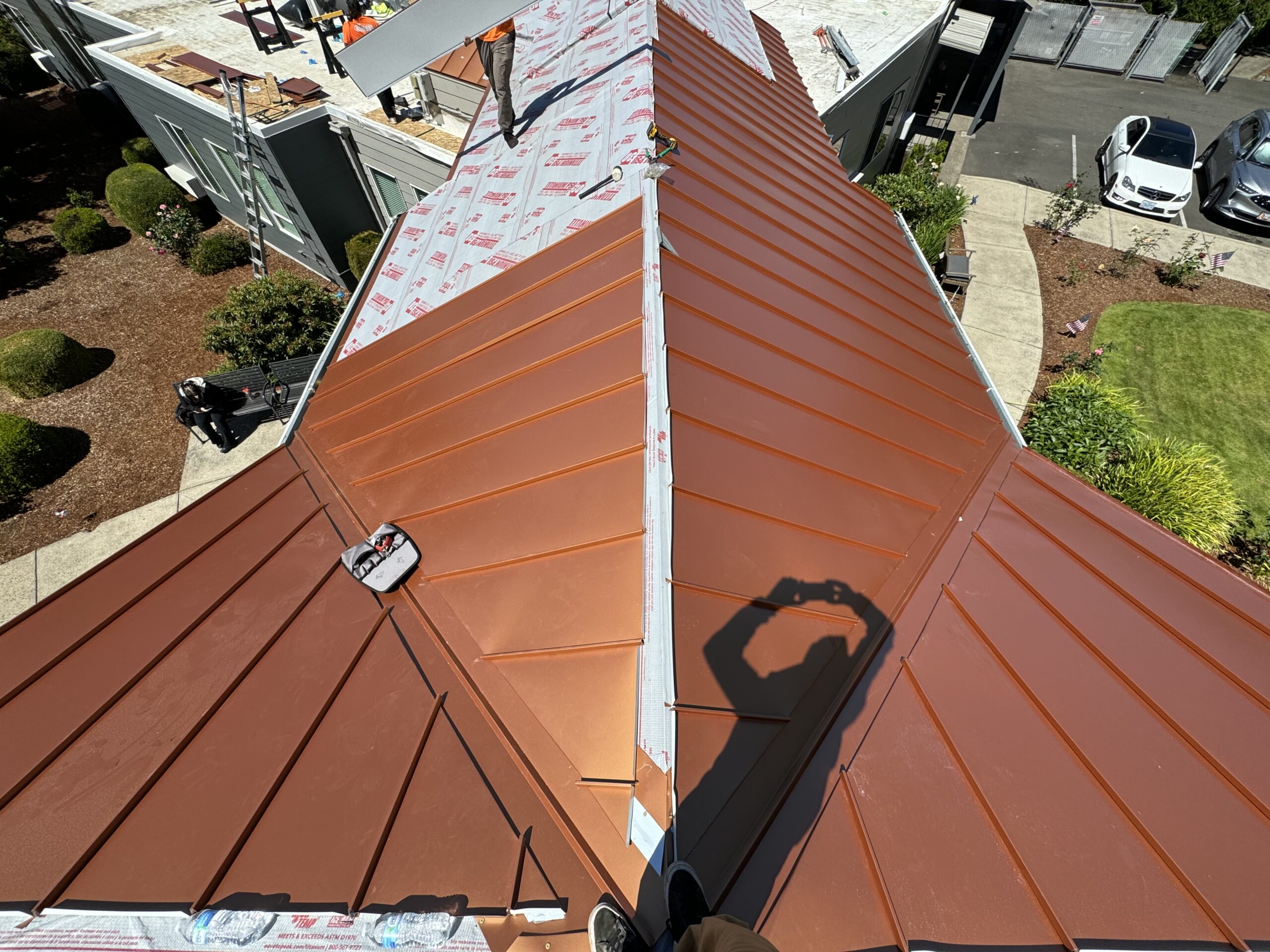 View of a newly installed brown metal roof on a residential building, with construction materials and tools visible. Nearby are green lawns and parked cars.