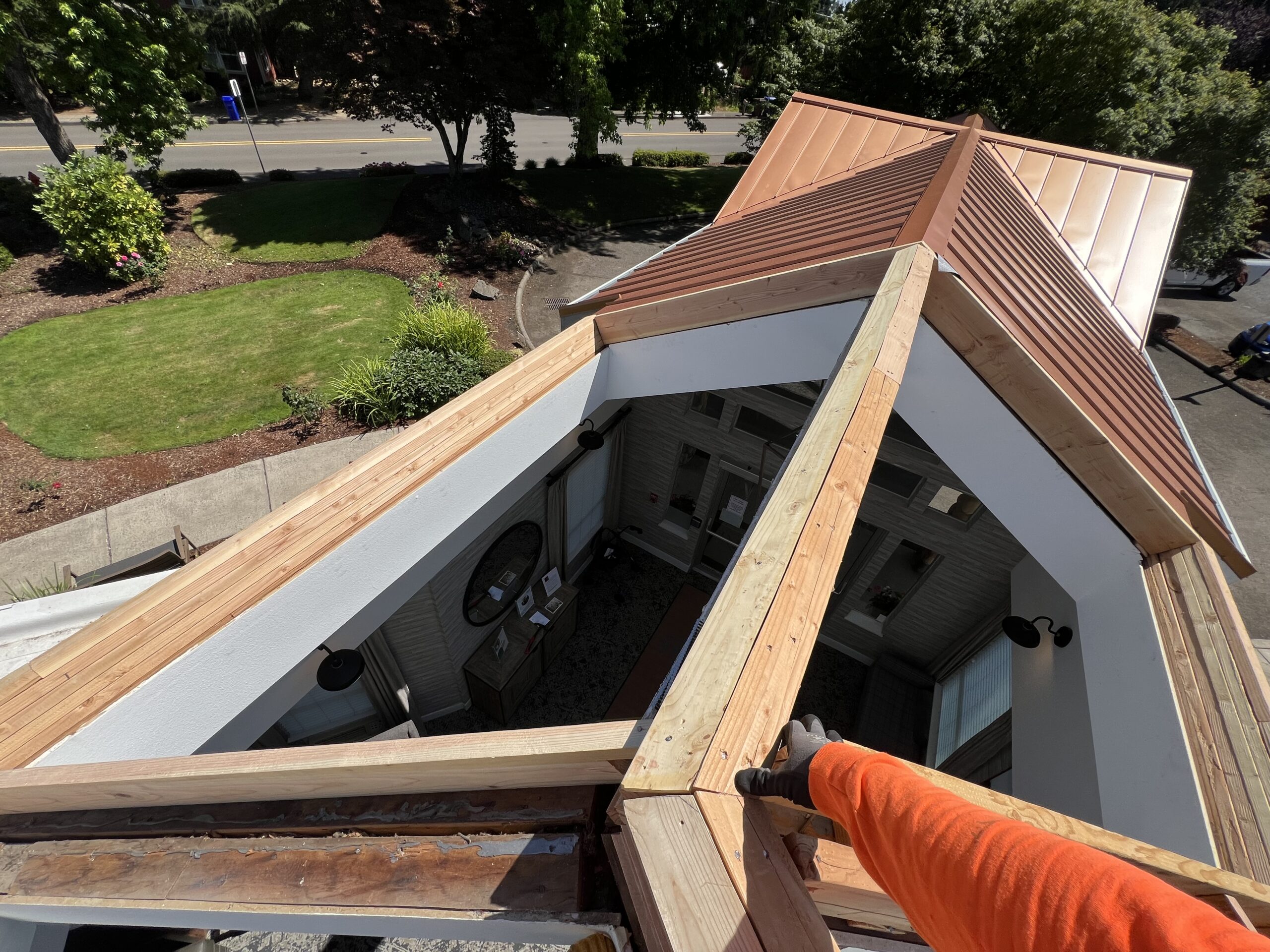 A person installing wooden beams on a roof with a view of the ground below, including a garden and walkway.