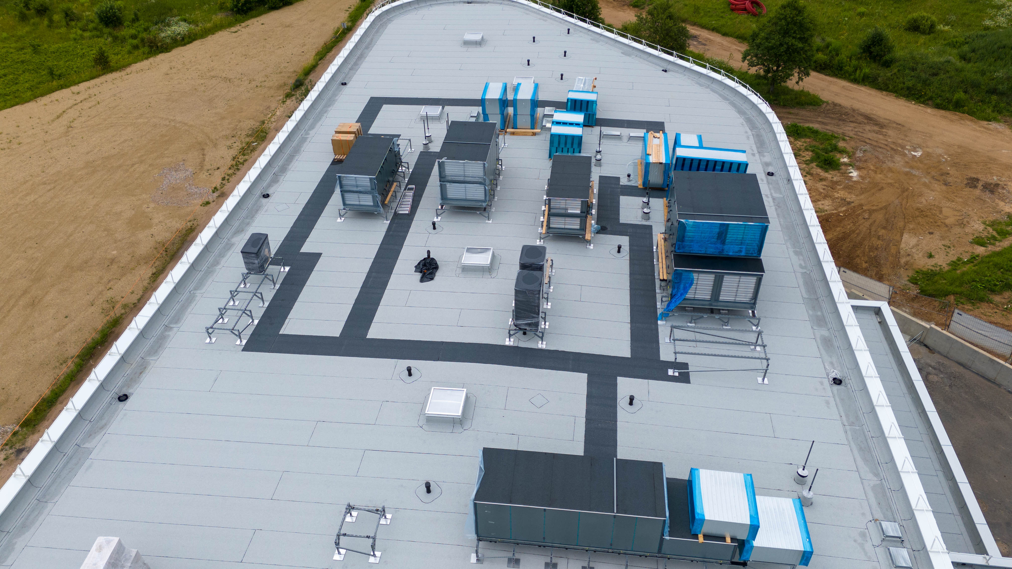 Aerial view of a flat rooftop with multiple HVAC units and vents, surrounded by greenery and construction areas.